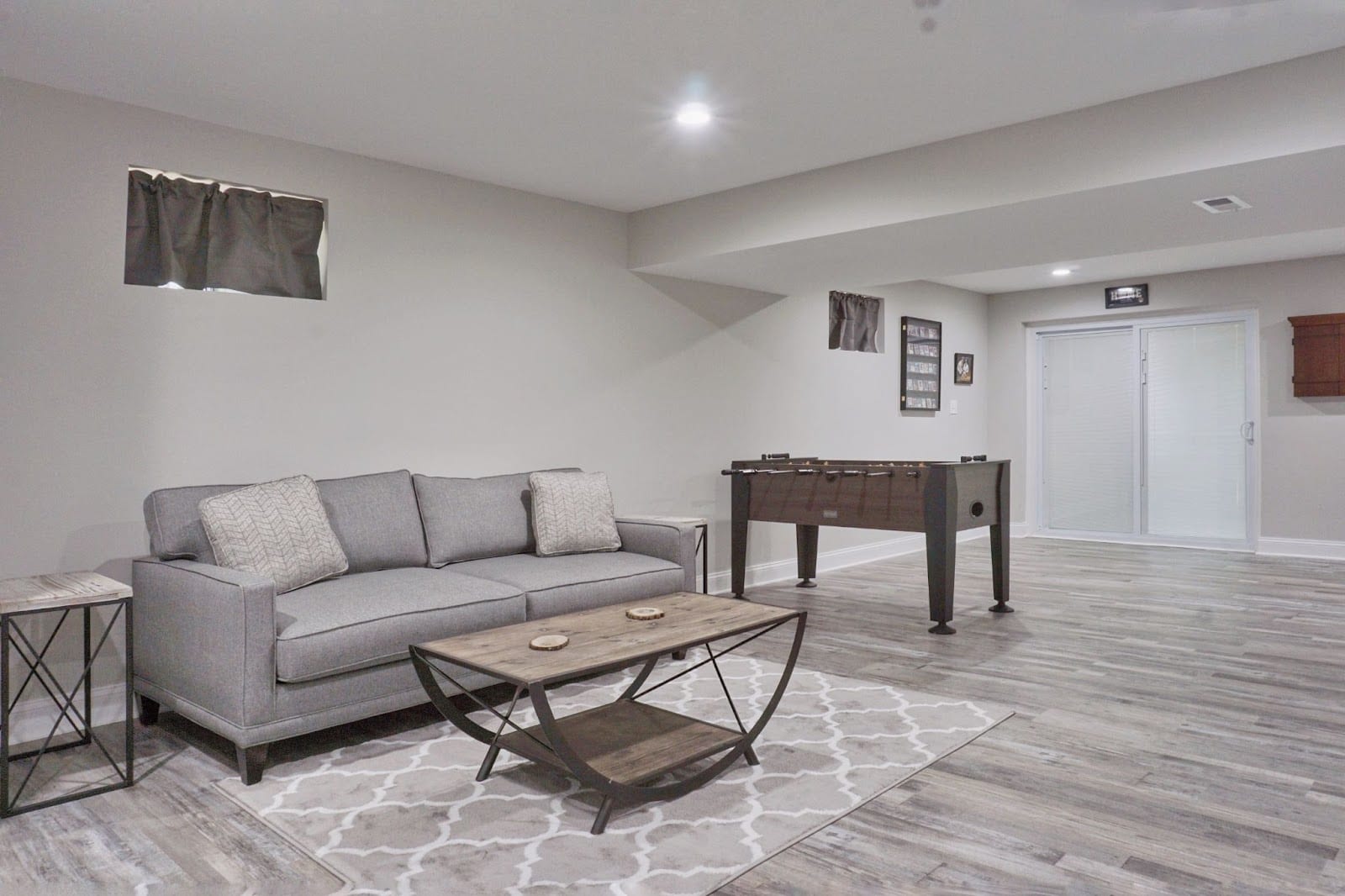 Modern basement room with a gray couch, coffee table, area rug, and foosball table on gray wood flooring.