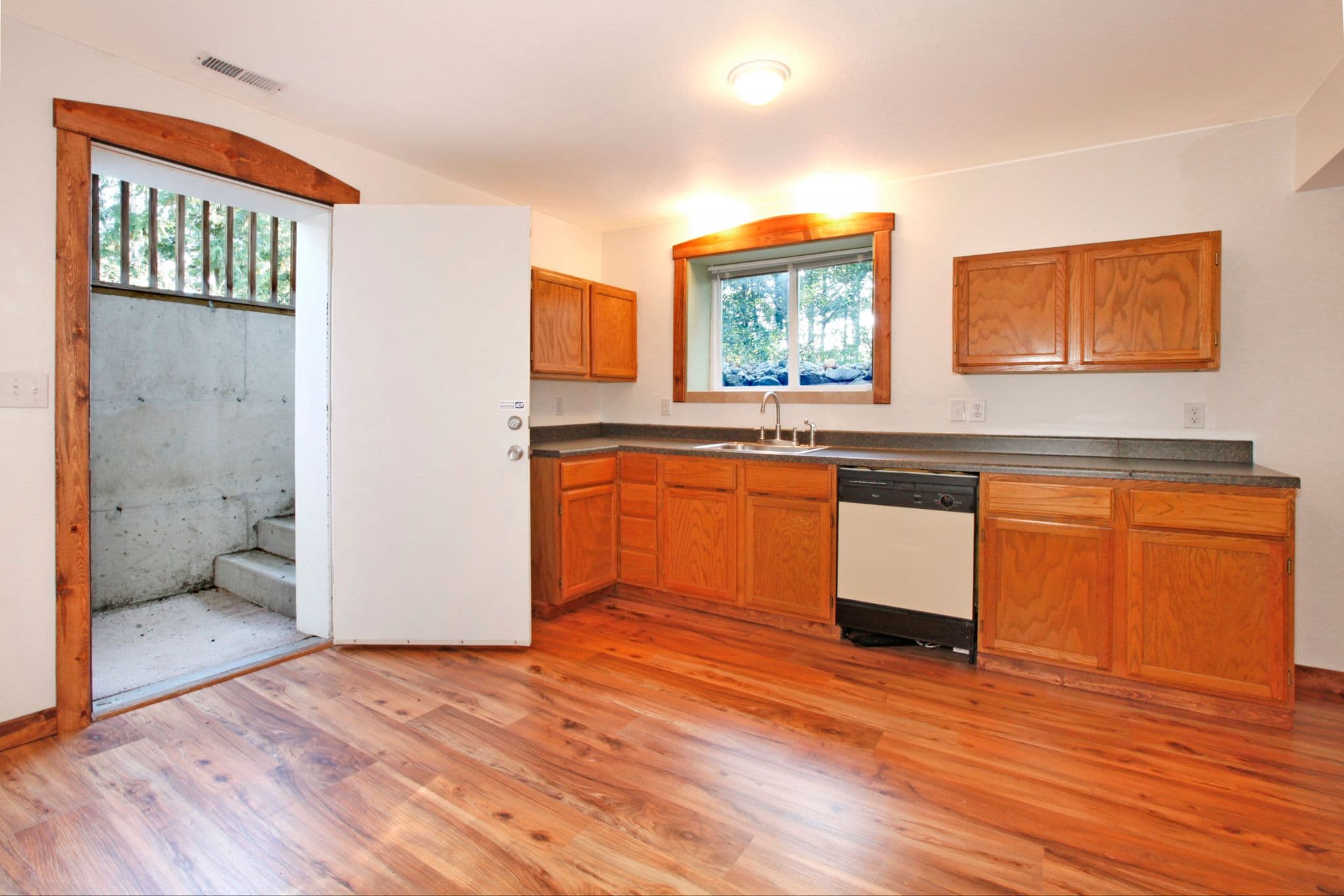 A basement kitchen with honey-oak cabinets and wood flooring next to an open door revealing concrete steps leading outside.
