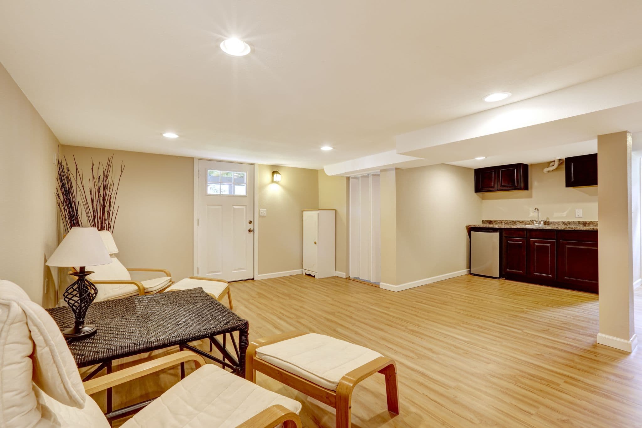 A finished walk-out basement with light wood flooring, a seating area with woven and wooden furniture, and a dark cabinet wet bar.