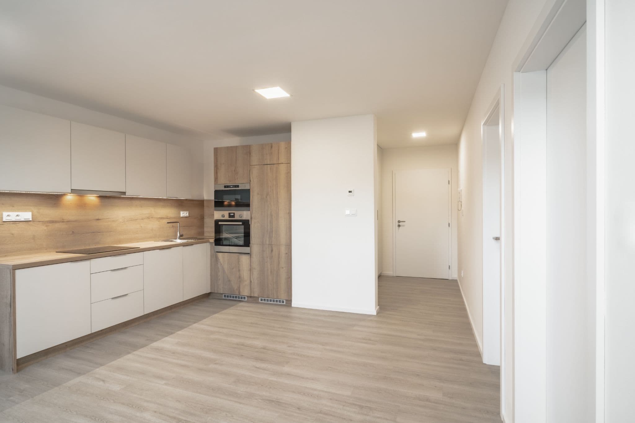 A modern minimalist kitchen with white and light wood cabinets, under-cabinet lighting, and an adjacent hallway.